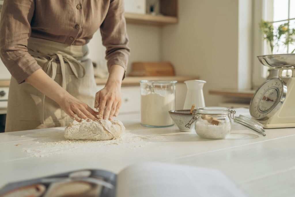 A person is kneading dough on a white kitchen countertop, surrounded by baking ingredients and tools.